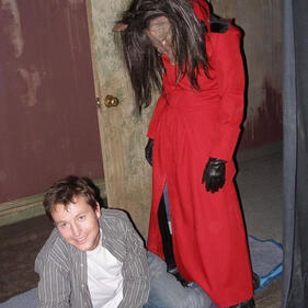 behind the scenes image from saw. leigh whannel is sitting on the floor, smiling and relaxed, and a person in a red hood and a pig mask is standing menacingly over him.